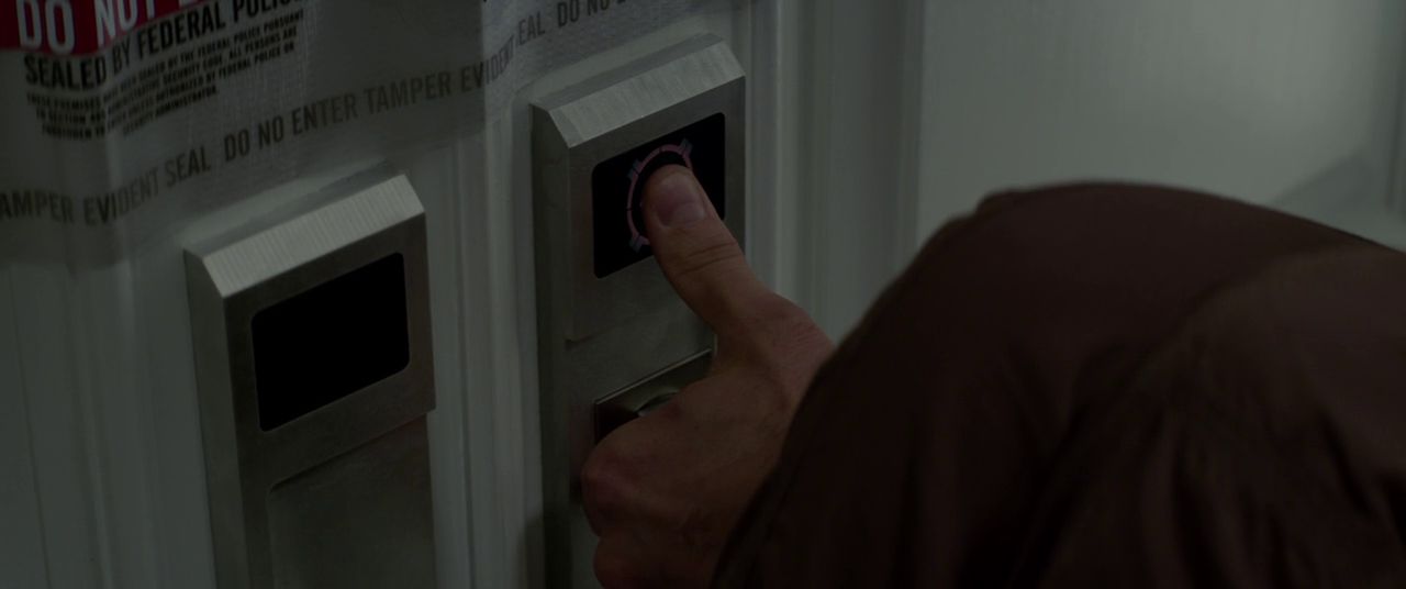 A thumb is pressed against a thumbprint scanner on what looks to be a very 80s tech panel on the wall